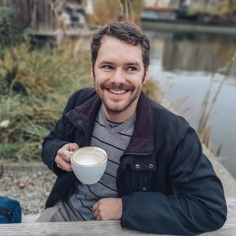 A smiling man holding a coffee cup sits outdoors by a pond with autumnal foliage in the background. He wears a jacket over a sweater, exuding a relaxed and joyful demeanor after completing his bathroom remodel RestoraCore