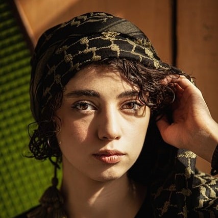 A close-up portrait of a young woman with curly hair, wearing a patterned scarf, her hand adjusting the scarf, set against a textured wooden background illuminated by warm light during a whole home remodel. RestoraCore